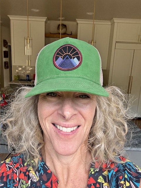 A person smiling, wearing a green cap with a colorful design, inside a kitchen.
