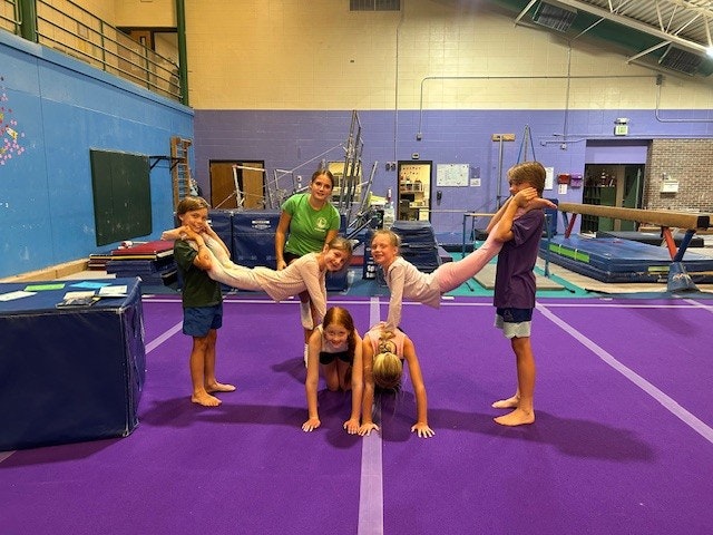 A group of children practicing gymnastics on a purple mat, forming human pyramids with a coach supervising.