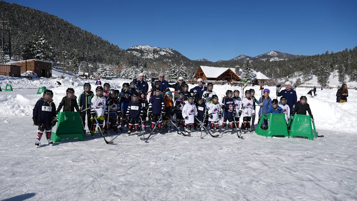 A group of children in hockey gear pose on a snowy outdoor ice rink, surrounded by trees and mountains under a clear blue sky.