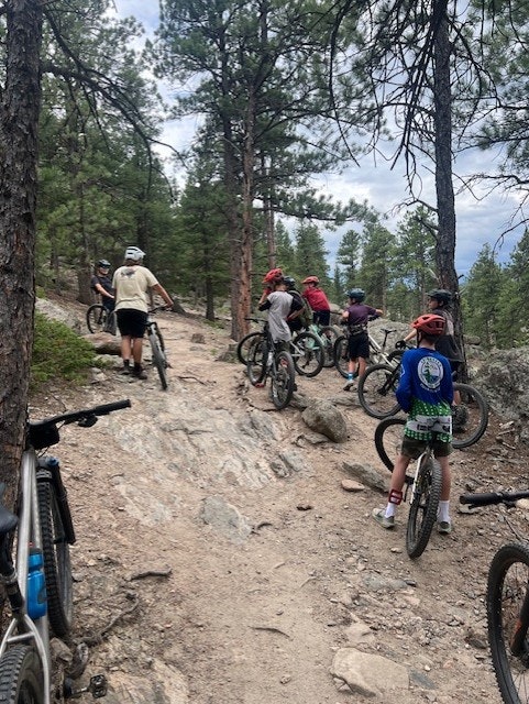 A group of kids in helmets and bikes pause on a rocky trail in a wooded area, surrounded by tall trees.