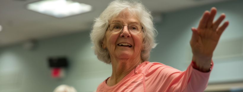An elderly woman with glasses, wearing a pink top, smiling and raising her hand in a bright indoor setting.