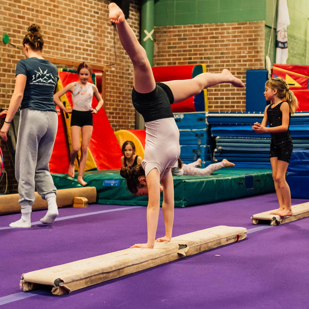A young gymnast performs a handstand on a balance beam in a gym, with others watching or practicing nearby.