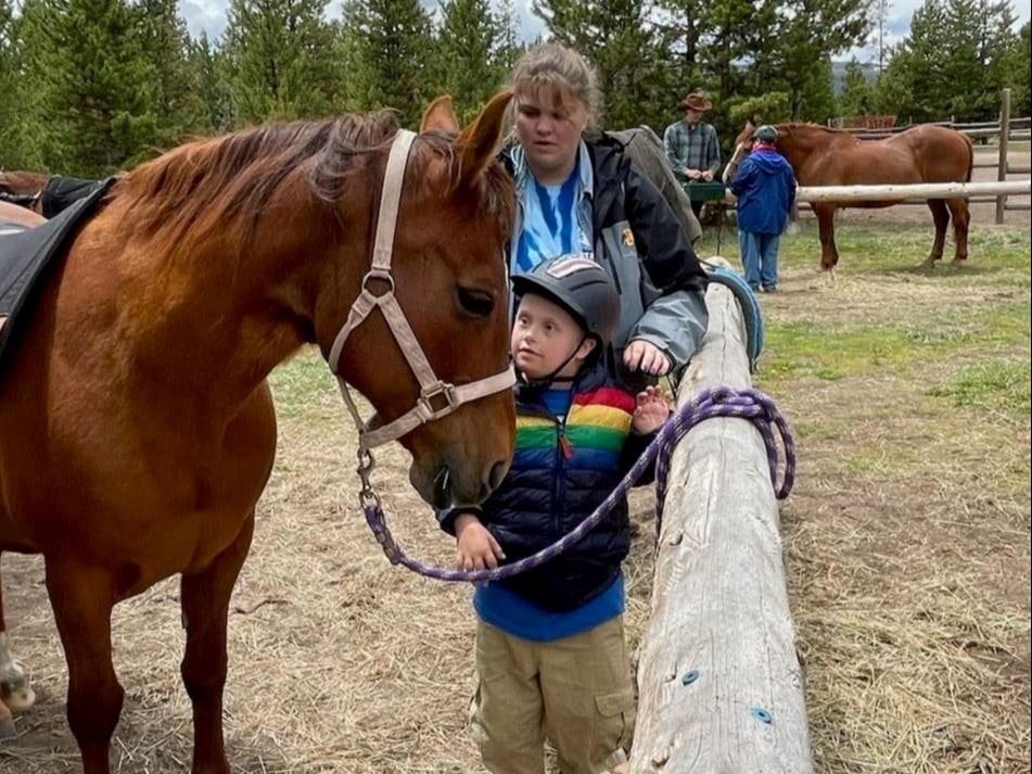 A child in a helmet stands by a horse, with a person behind them, and others near horses in the background.