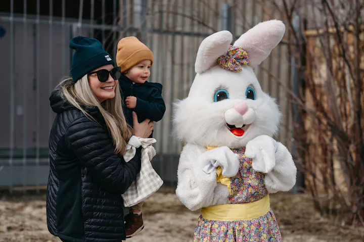 A woman holds a child while posing with a person in a bunny costume, set outdoors with trees in the background.