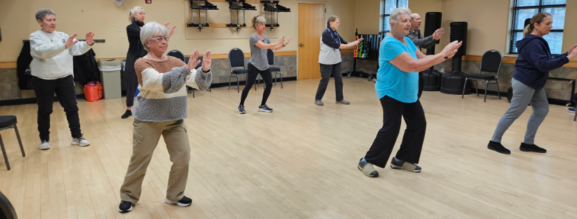 A group of people practicing tai chi in a room with wooden floors and chairs against the walls.