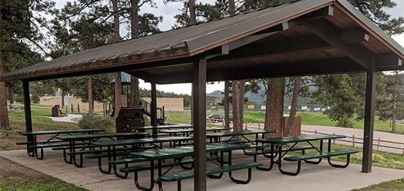 A covered picnic area with tables and a barbecue grill in a park setting.