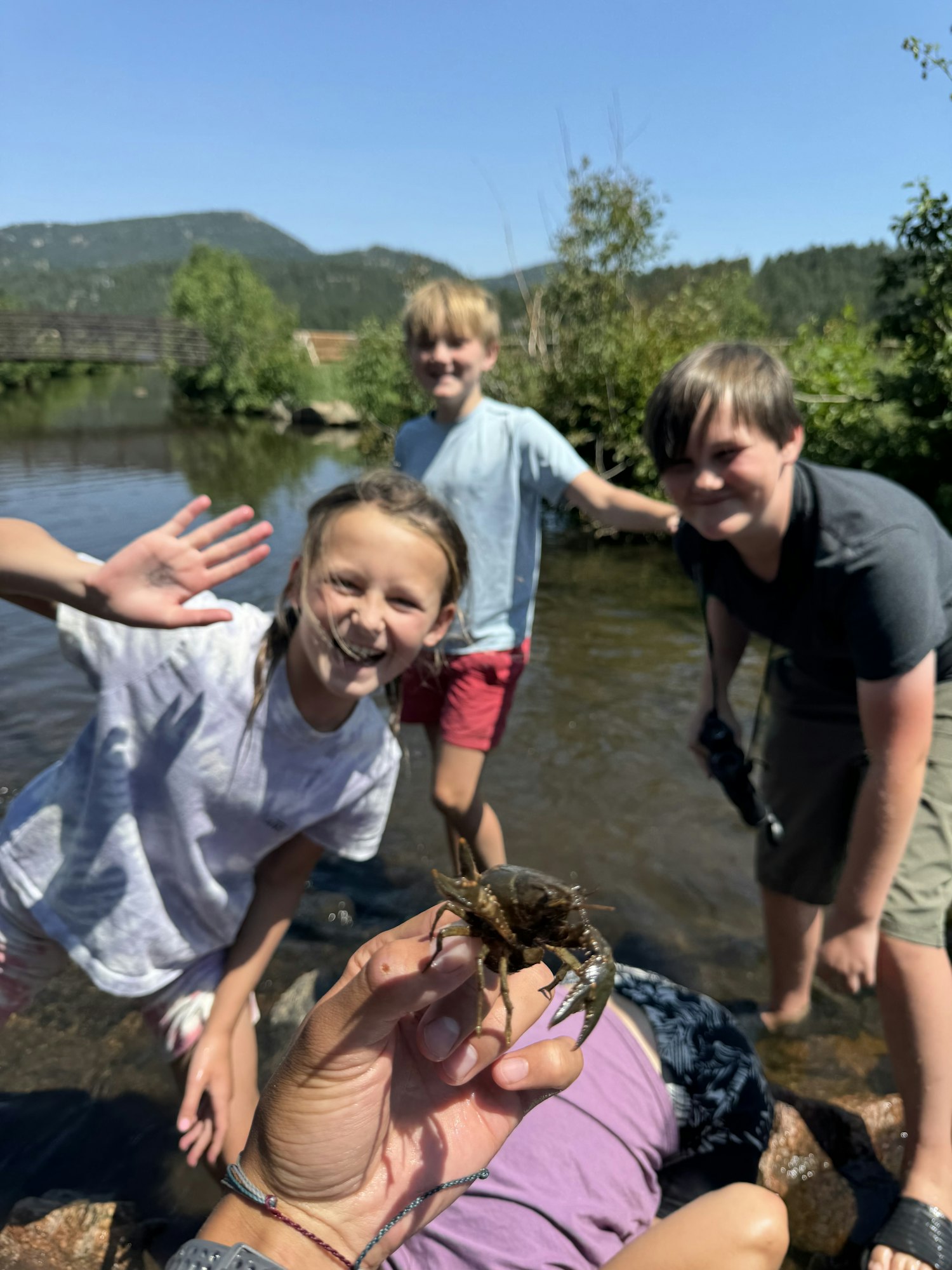 A group of kids is outdoors by a river. One holds a crayfish. The background shows trees and mountains.