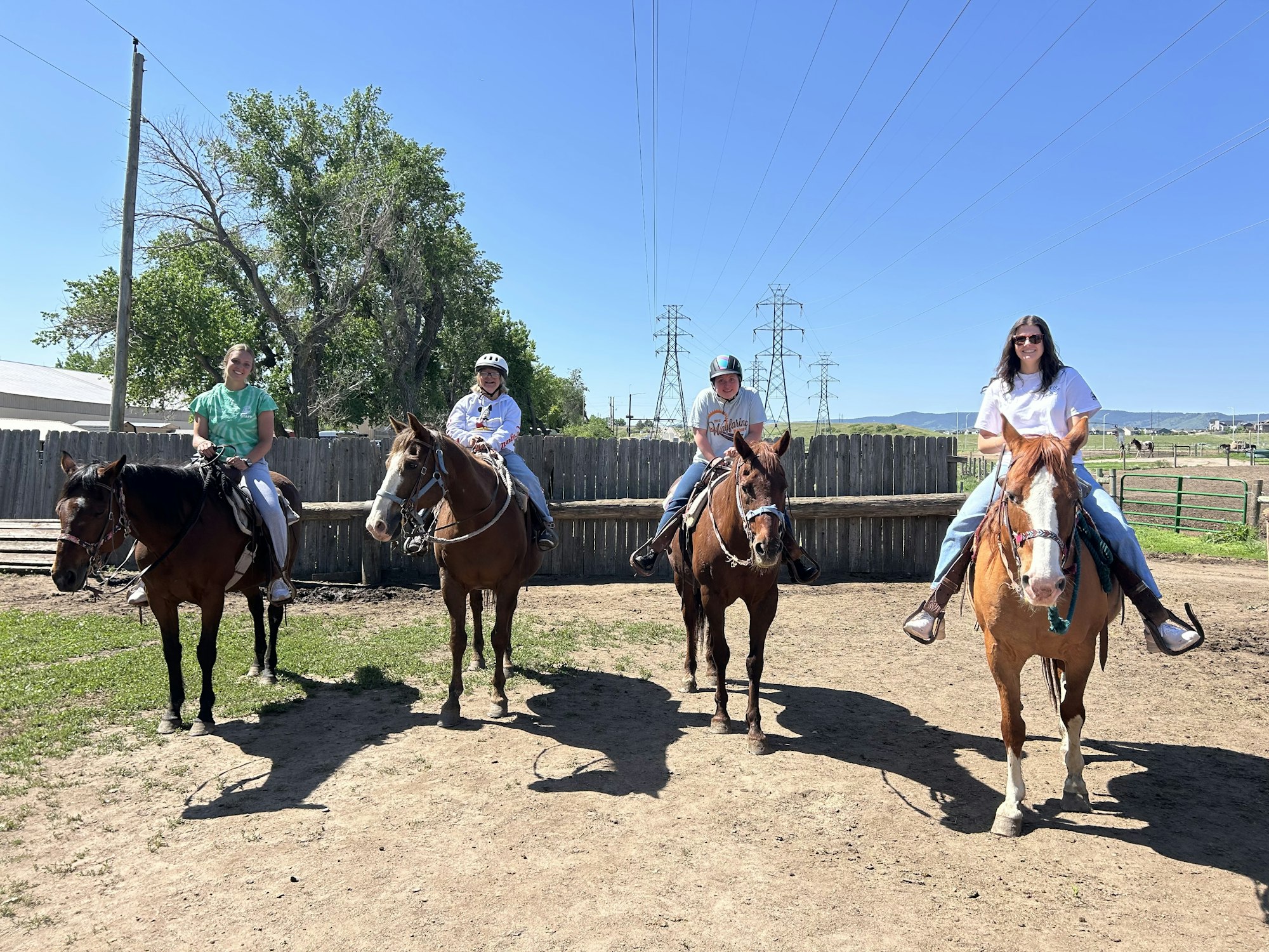 Four people riding horses in an outdoor setting with a wooden fence and clear sky.