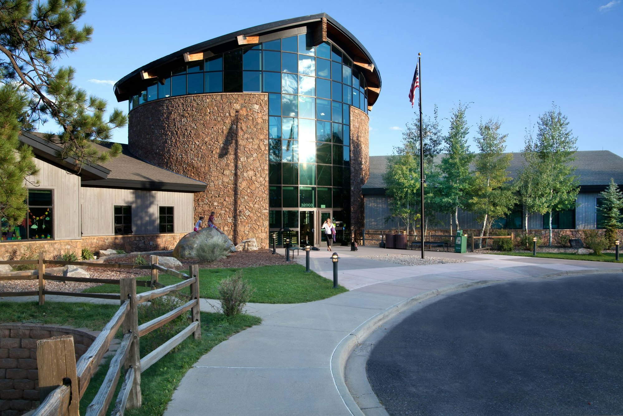 Modern building with glass and stone facade, surrounded by trees under a blue sky, with a flagpole and people nearby.