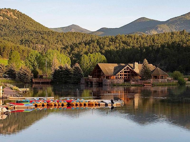 Lakeside log cabin, dock with colorful kayaks, forested hills, serene water reflection.