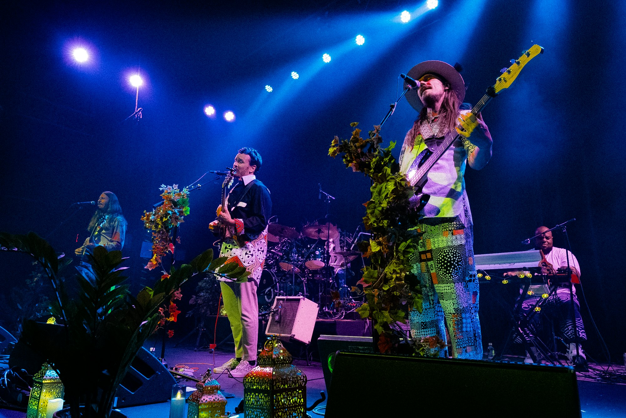 A vibrant live music performance featuring a band with colorful outfits, plants, and atmospheric lighting.