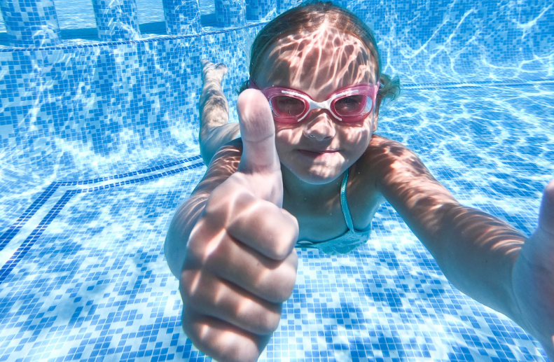 Child underwater in a pool, wearing pink goggles, giving a thumbs up with a bright smile and blue mosaic tiles in the background.