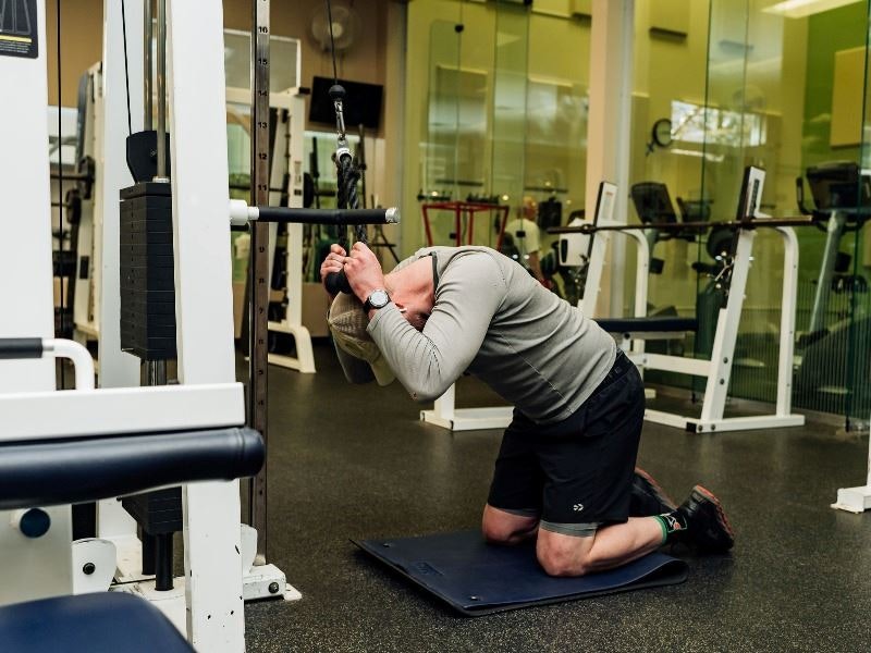 A person is stretching or exercising on a mat in a gym with various fitness equipment around.