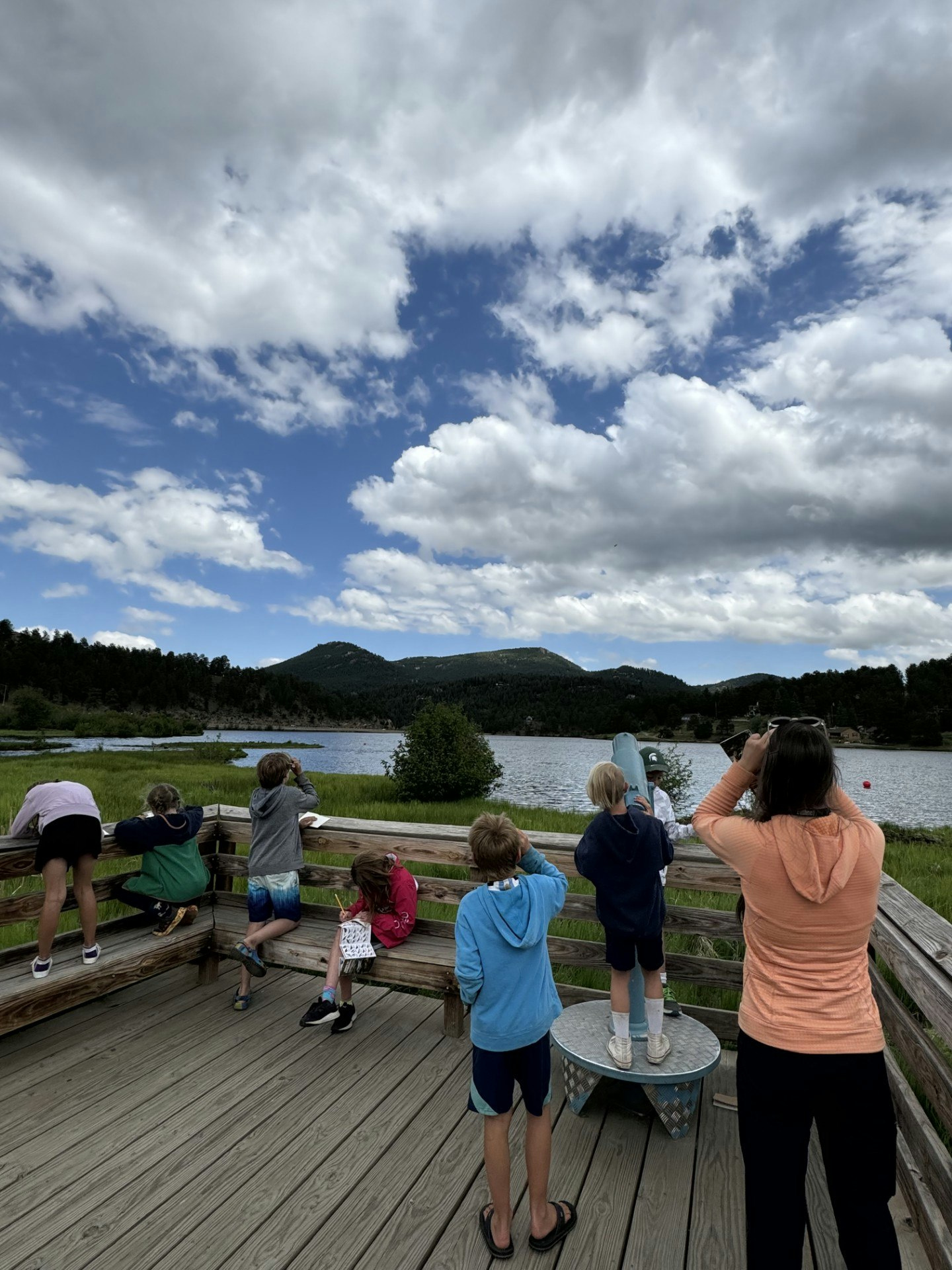 A group of people on a wooden deck observing a scenic lake view with mountains and cloudy sky.