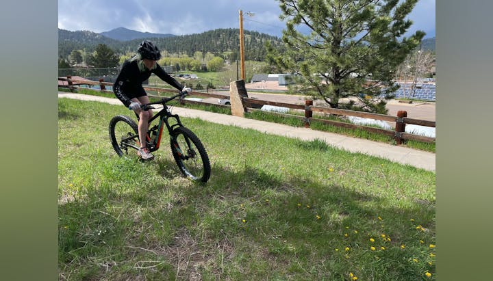 A person in cycling gear rides a mountain bike on a grassy hill, with trees and mountains in the background.