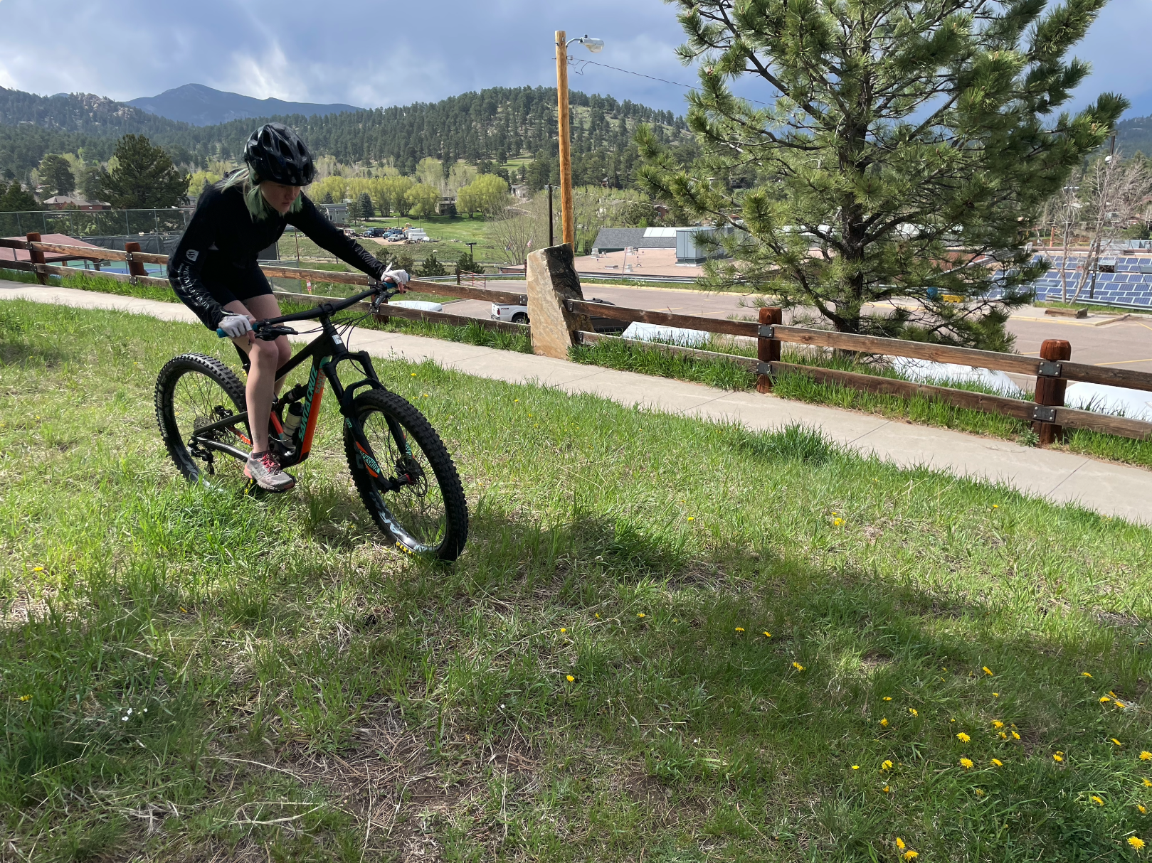 A person in cycling gear rides a mountain bike on a grassy hill, with trees and mountains in the background.