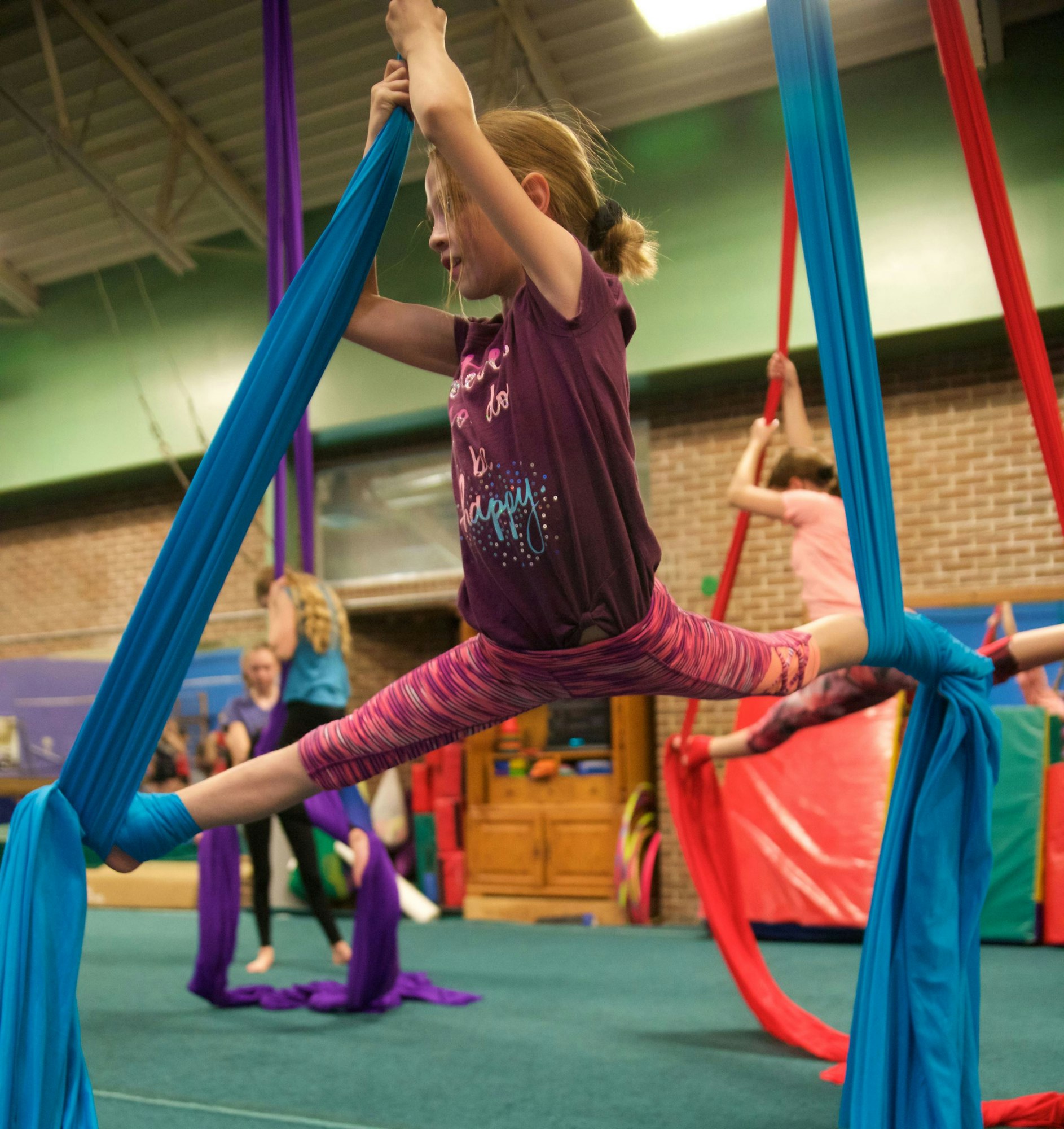 A young girl practices aerial silk acrobatics in a gym, with colorful fabrics and participants in the background.