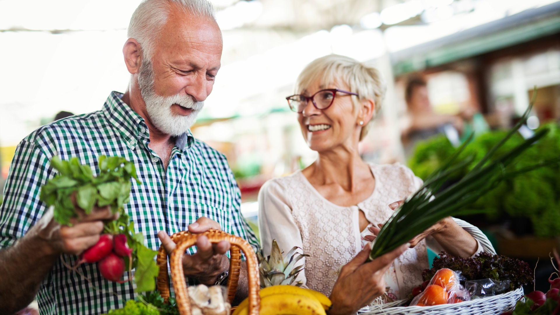 An elderly couple happily shopping for fresh vegetables and fruit at a market.
