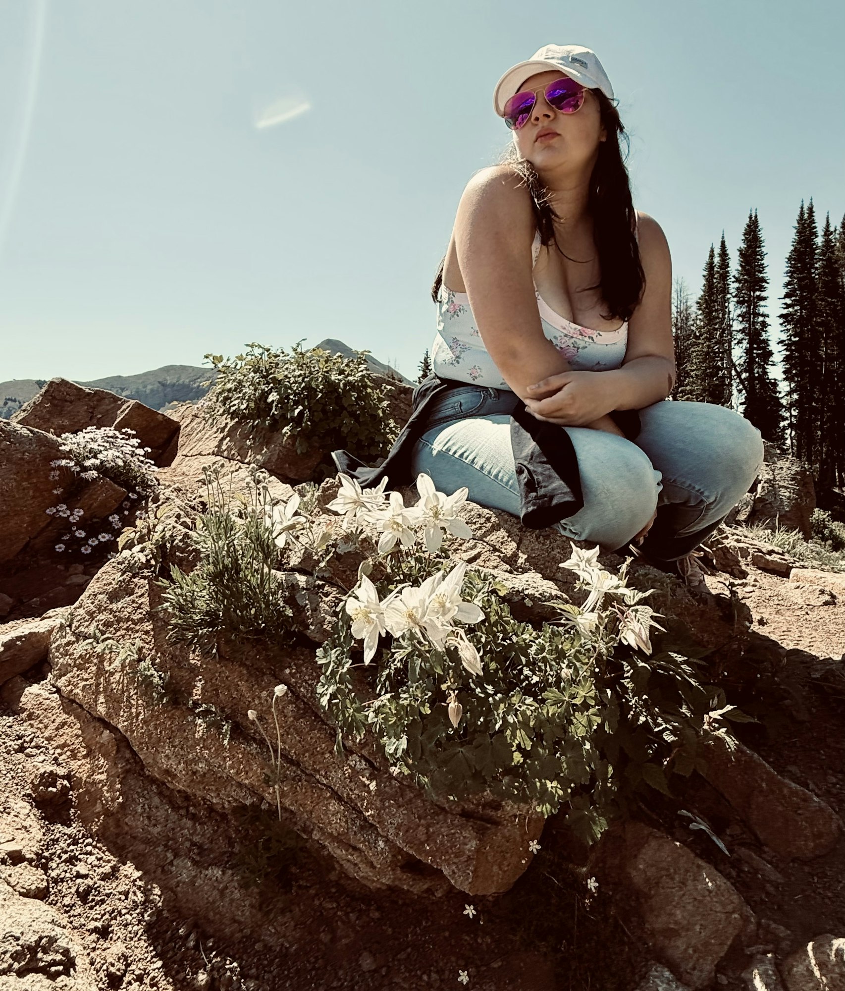 A person poses on a rock among flowers, wearing sunglasses and a cap, with mountains and trees in the background.
