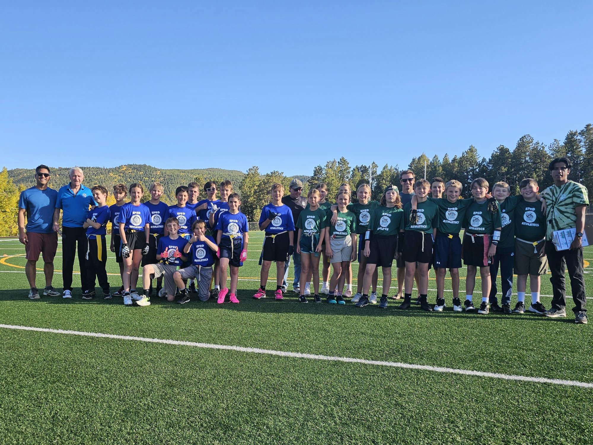 A group of kids in jerseys poses on a sports field, with adults nearby, showing a moment from a team event or league gathering.