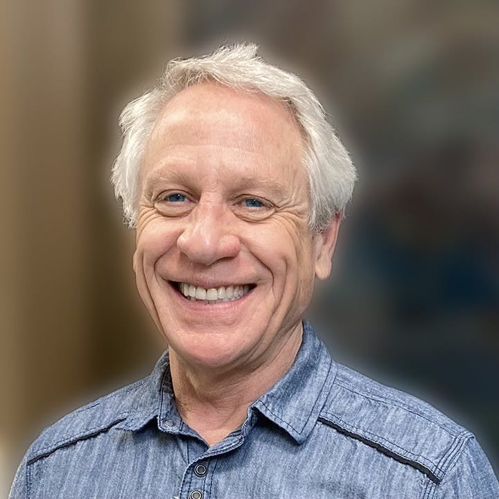 The image shows a smiling man with light hair, wearing a blue collared shirt. The background appears blurred.