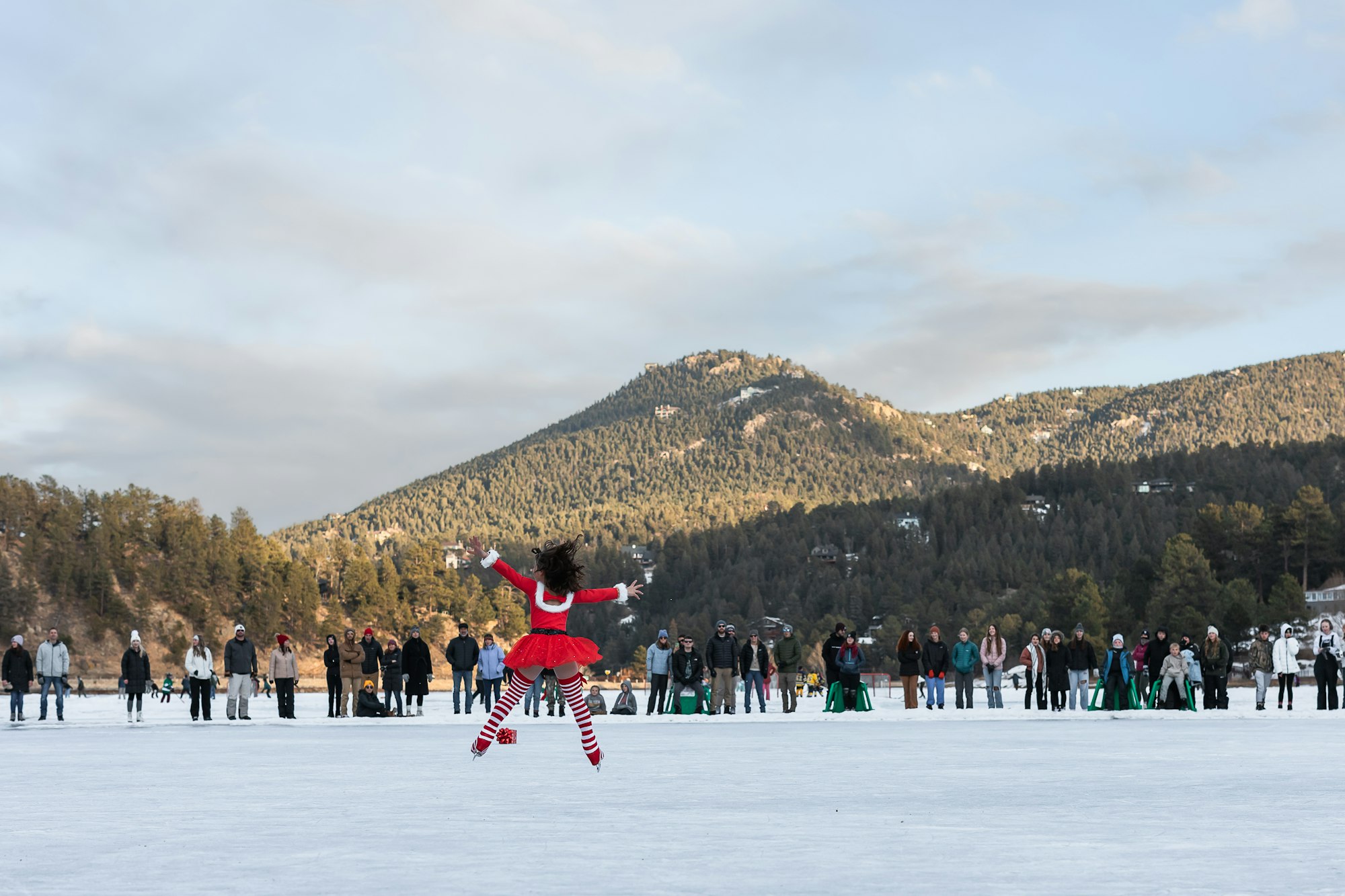 A person in a festive outfit spins on ice, observed by a crowd against a mountainous backdrop.