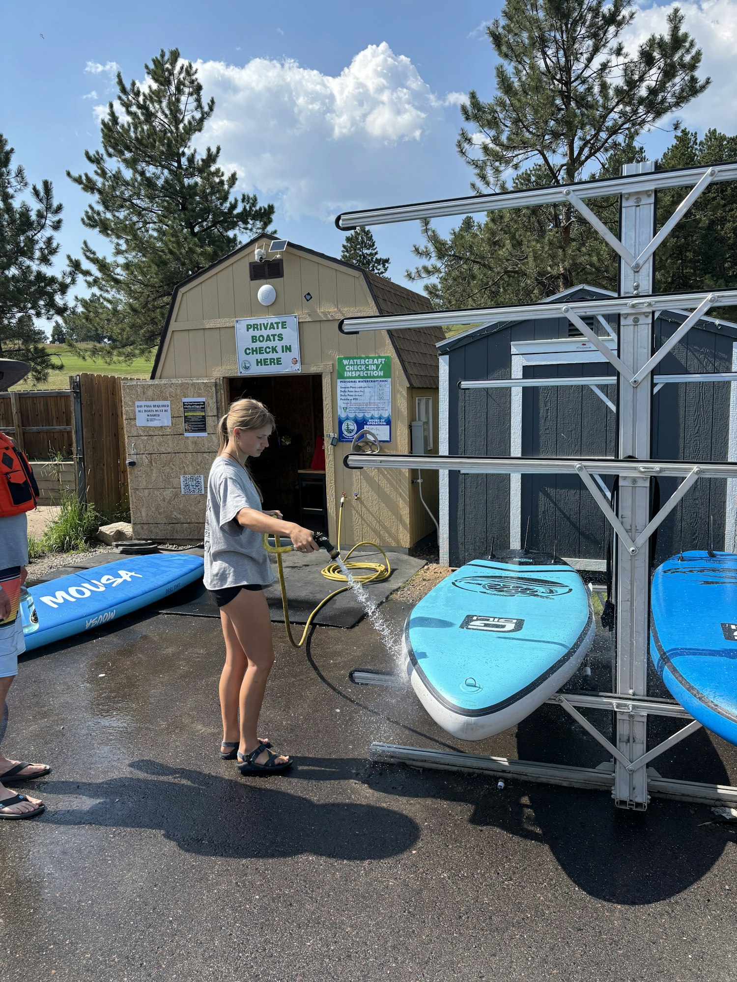 Person washing blue paddleboards with a hose near a building labeled "PRIVATE BOATS CHECK IN HERE."