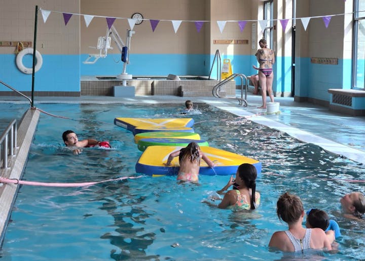 Indoor pool with kids playing on floating mats, supervised by adults.