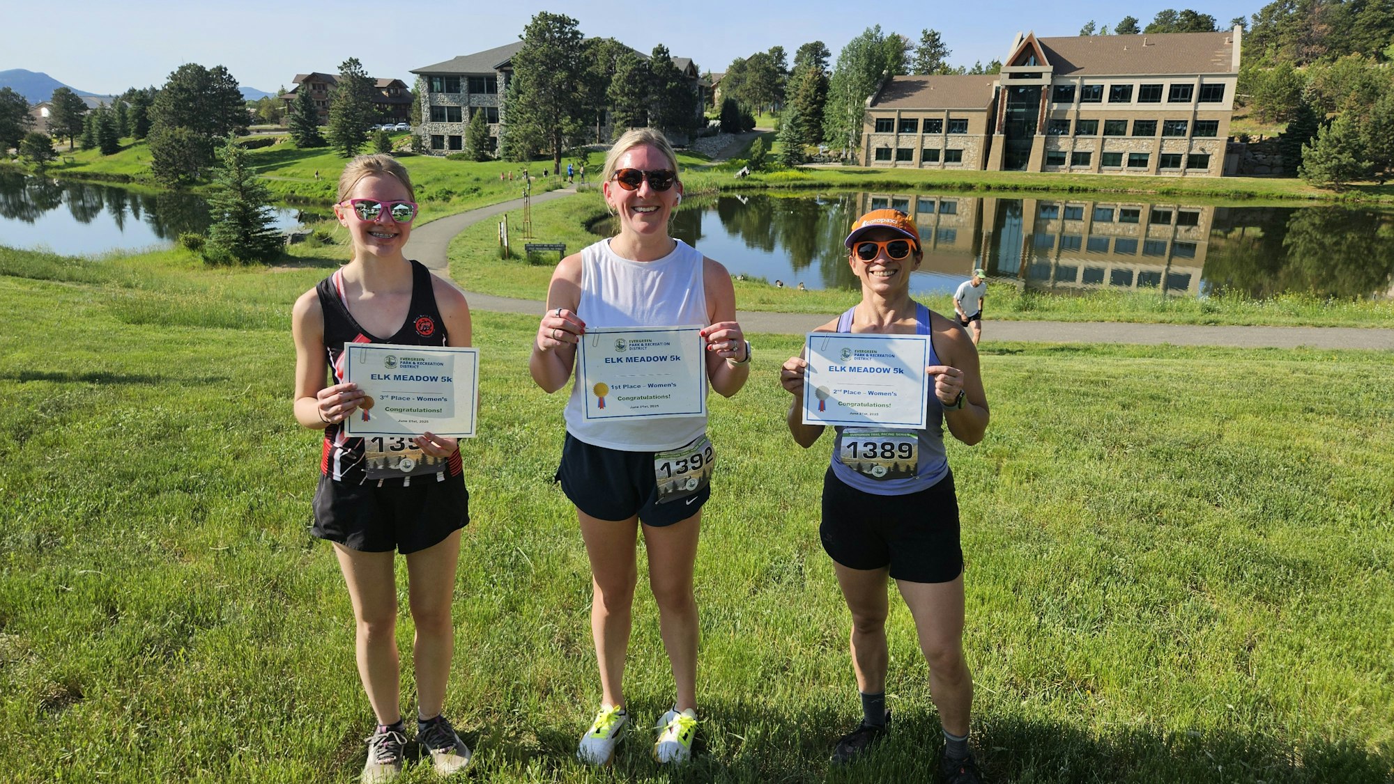 Three people holding certificates at an outdoor event by a lake.