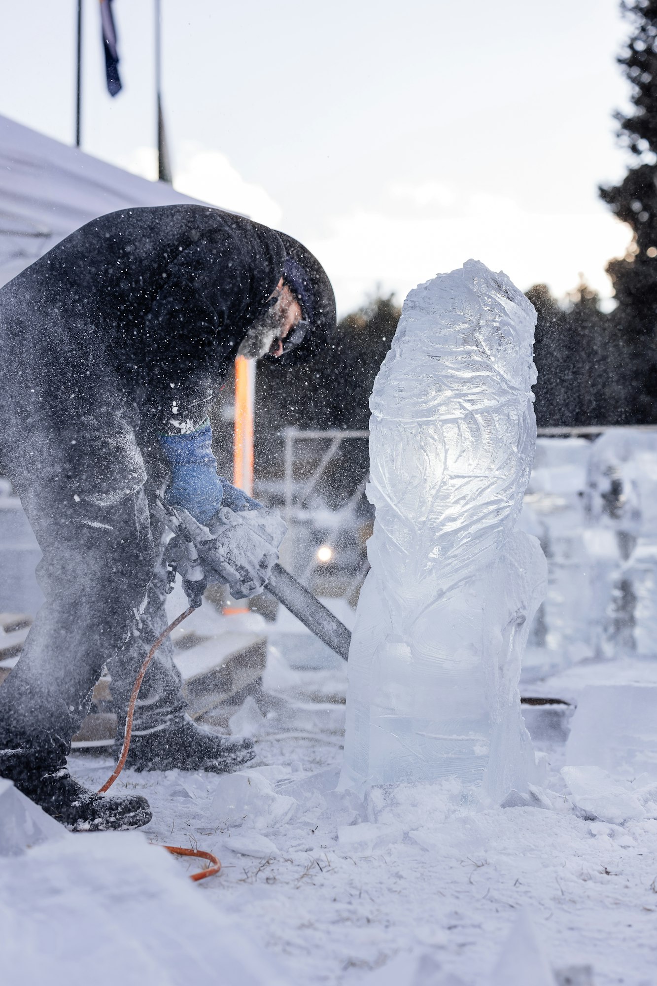 An artist wearing winter clothing sculpts ice with a chainsaw, surrounded by snow and ice shards.