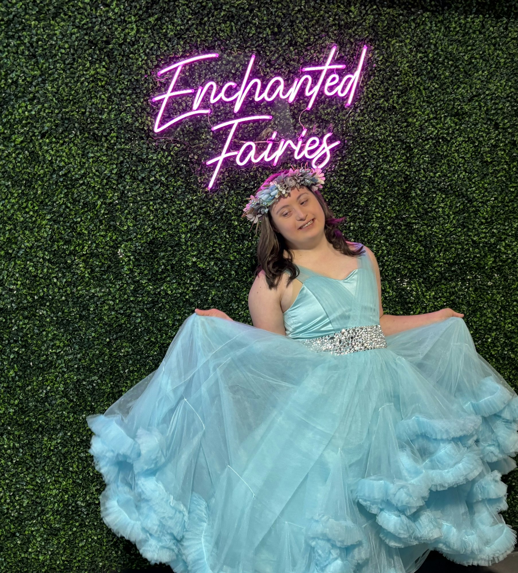 A person in a blue dress and floral crown stands in front of a neon sign reading "Enchanted Fairies" on a leafy backdrop.
