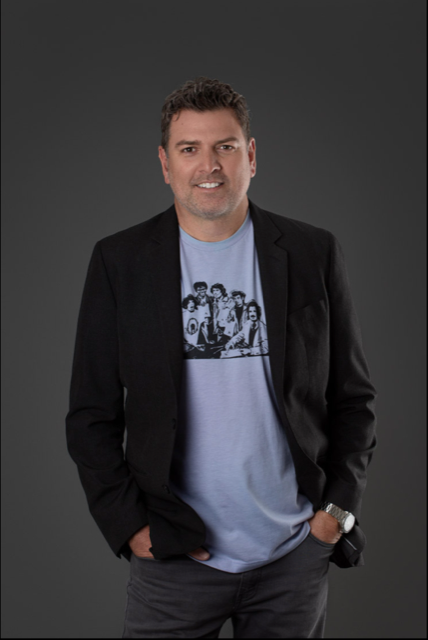 A man in a blazer over a graphic T-shirt stands against a dark background, smiling with hands in pockets.