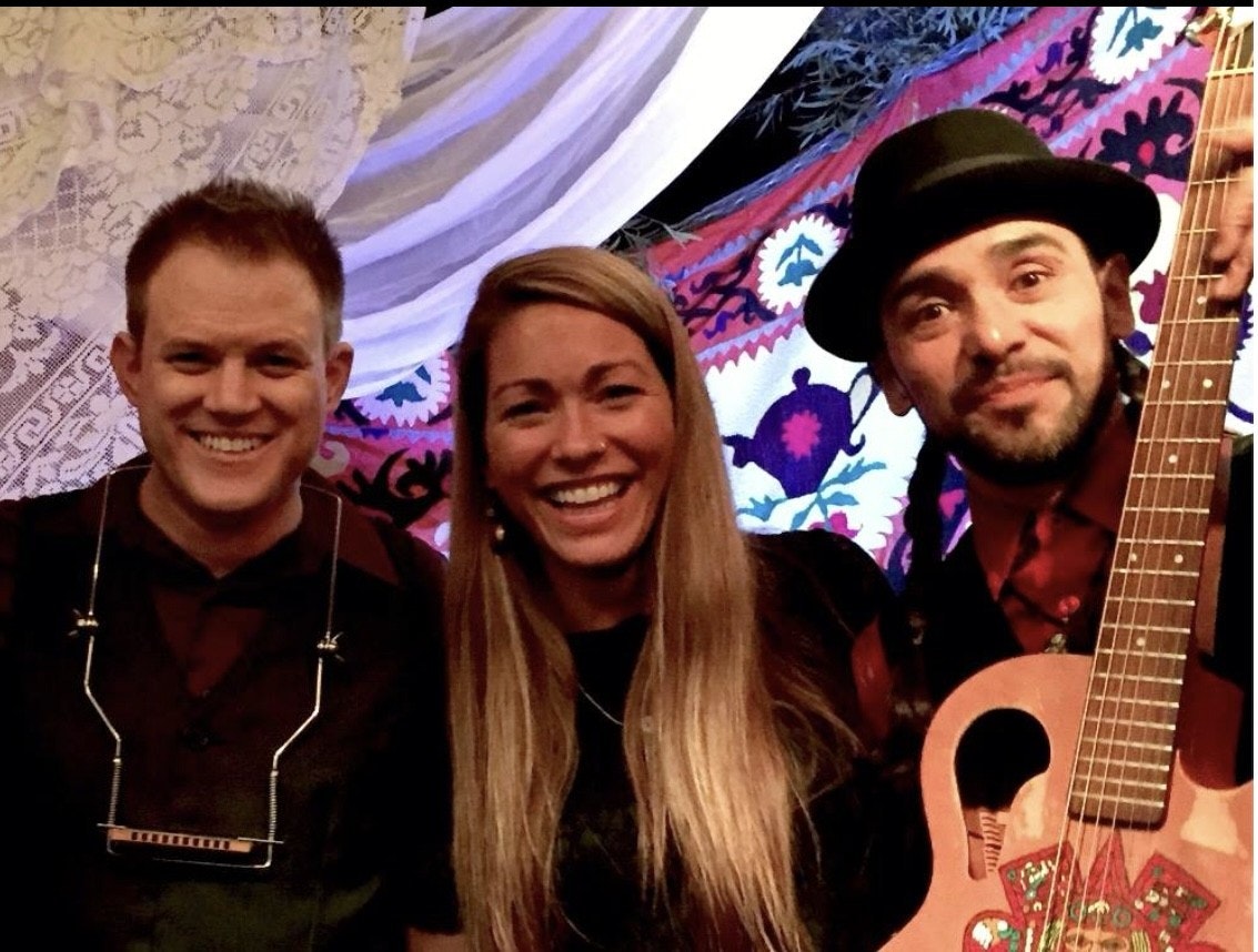 Three people smiling; one holds a guitar, another has a harmonica. Colorful tapestry backdrop.