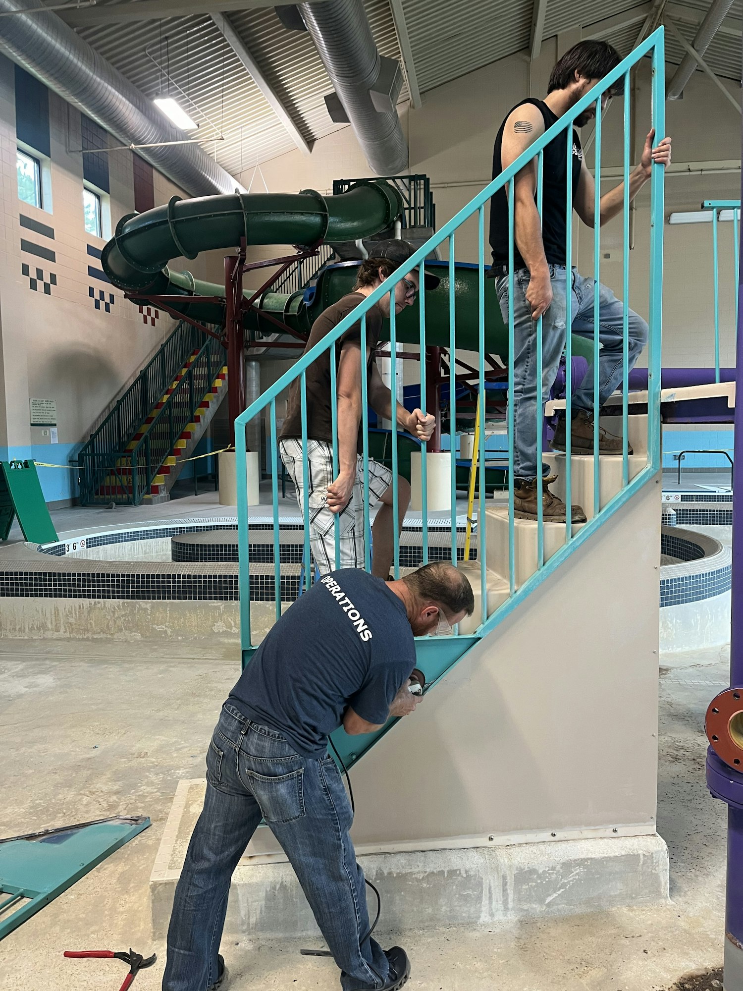 Three people are working on a light blue railing, with a green slide in the background of an indoor water facility.