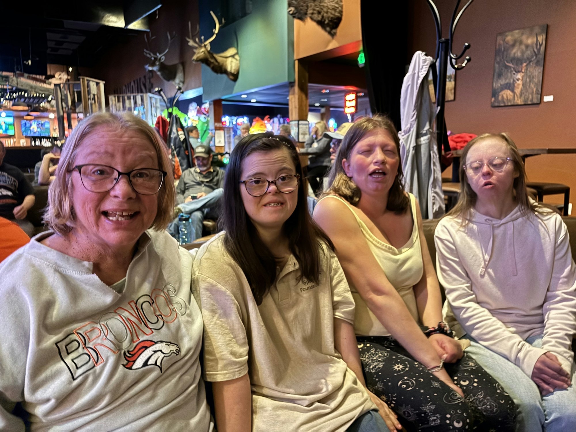 Four people sitting together indoors with a Broncos shirt visible. The background features mounted deer heads and colorful lights.