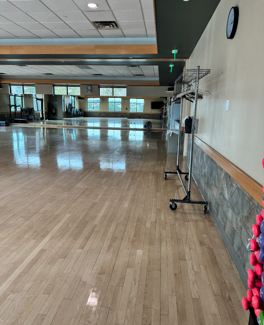 Empty fitness studio with hardwood floors, mirrors, exercise equipment, clocks, and a coat rack.
