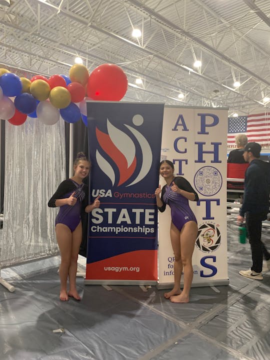 Two gymnasts in purple outfits pose by a USA Gymnastics State Championships sign, with balloons and American flags in the background.