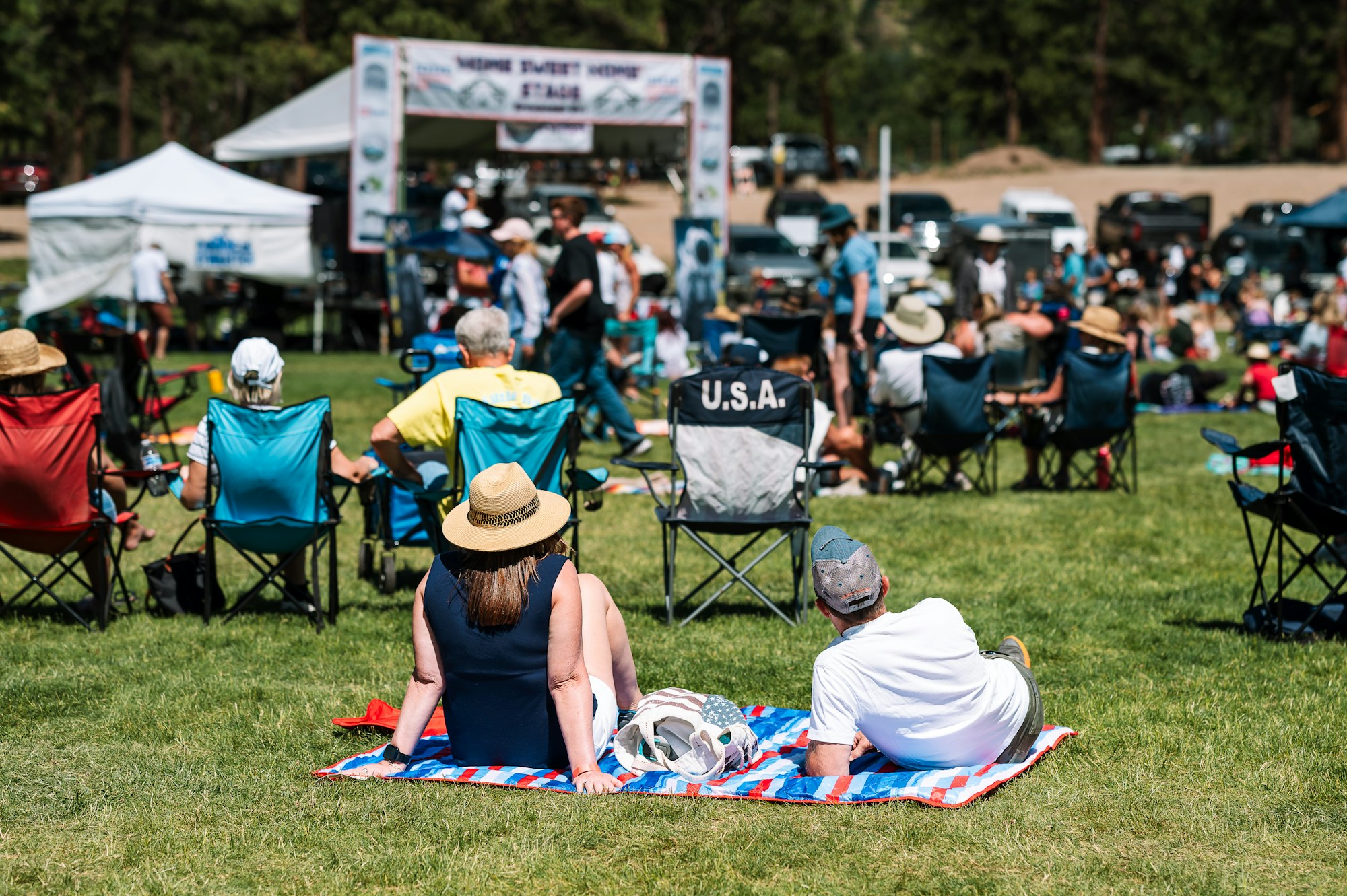 People sitting on chairs and blankets at an outdoor event or festival in a park.