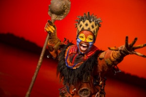 A performer in colorful makeup and costume holds a staff on a stage with a red background.