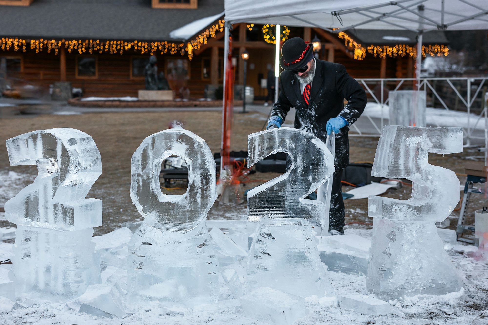 Man carving ice sculptures of the numbers "2025" under a canopy, with festive lights in the background.