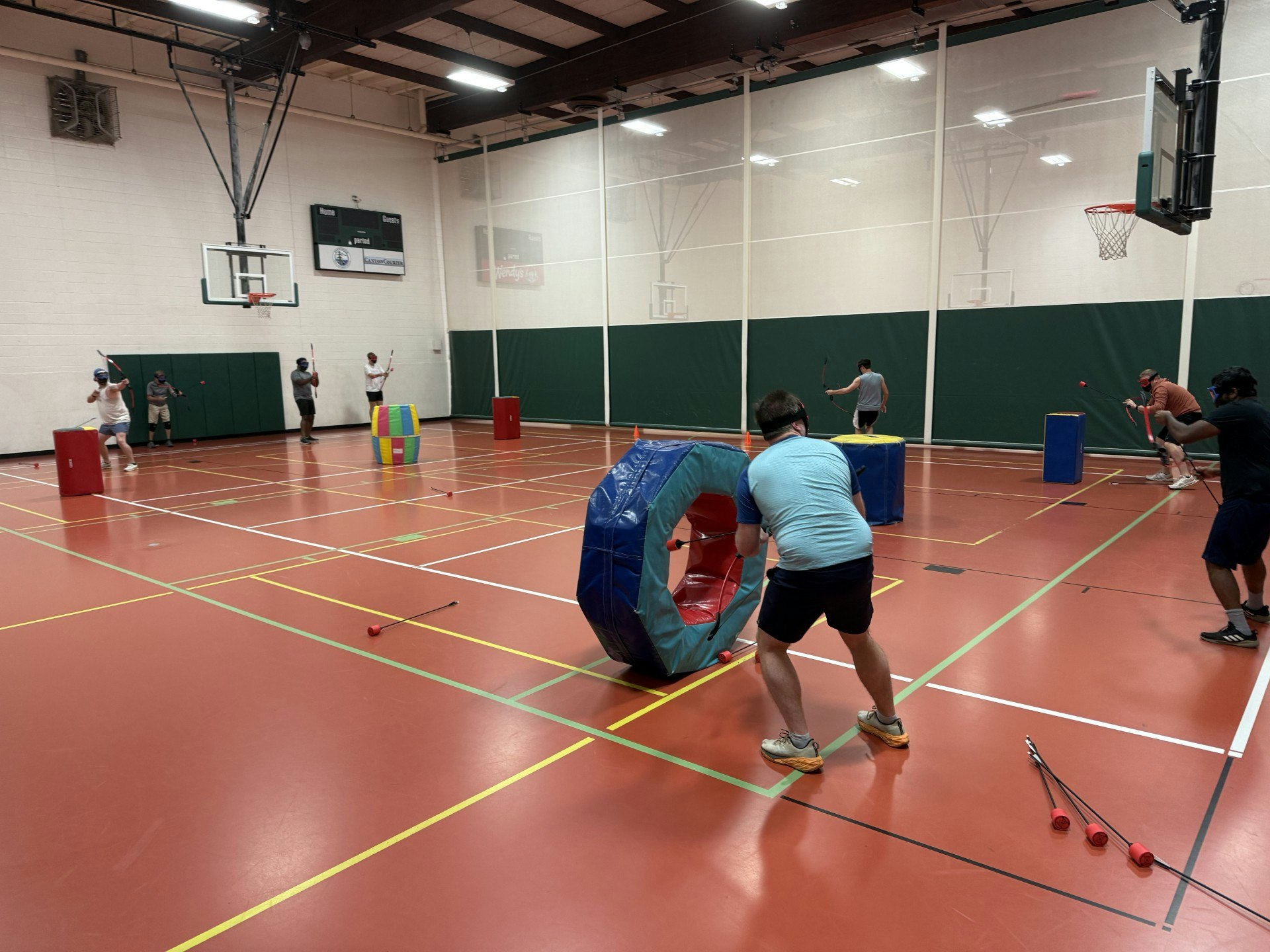 People playing archery tag in a gym, using foam-tipped arrows and inflatable obstacles.