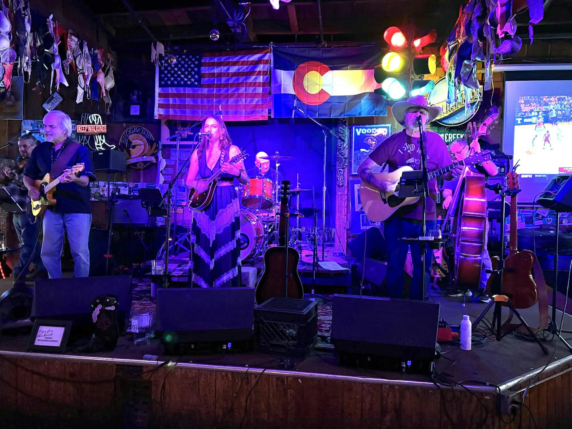 A band performing on stage with instruments, colorful lights, and wall decorations, including flags and bras.