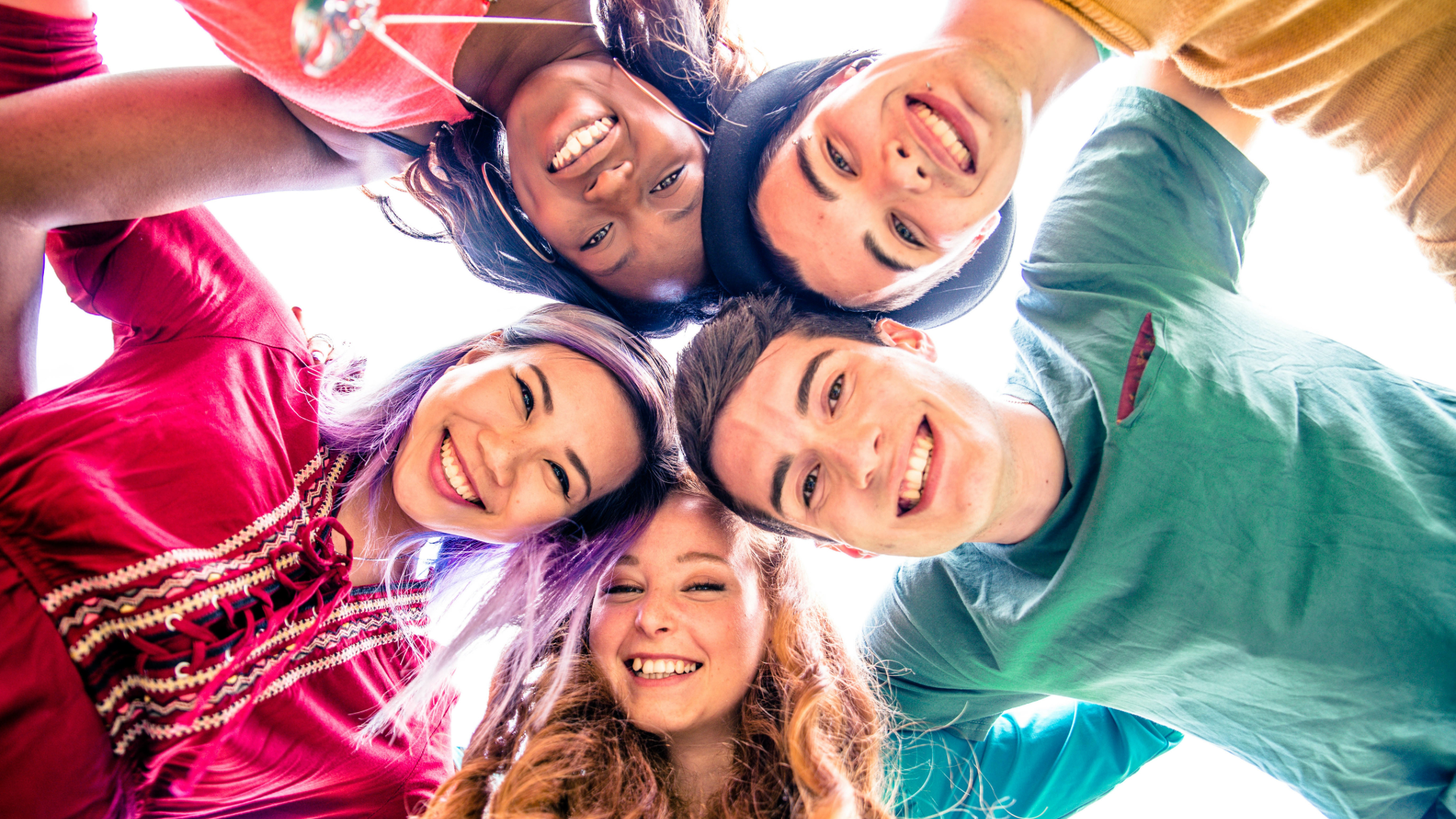 Five people looking down at the camera, smiling and embracing, in bright clothes.