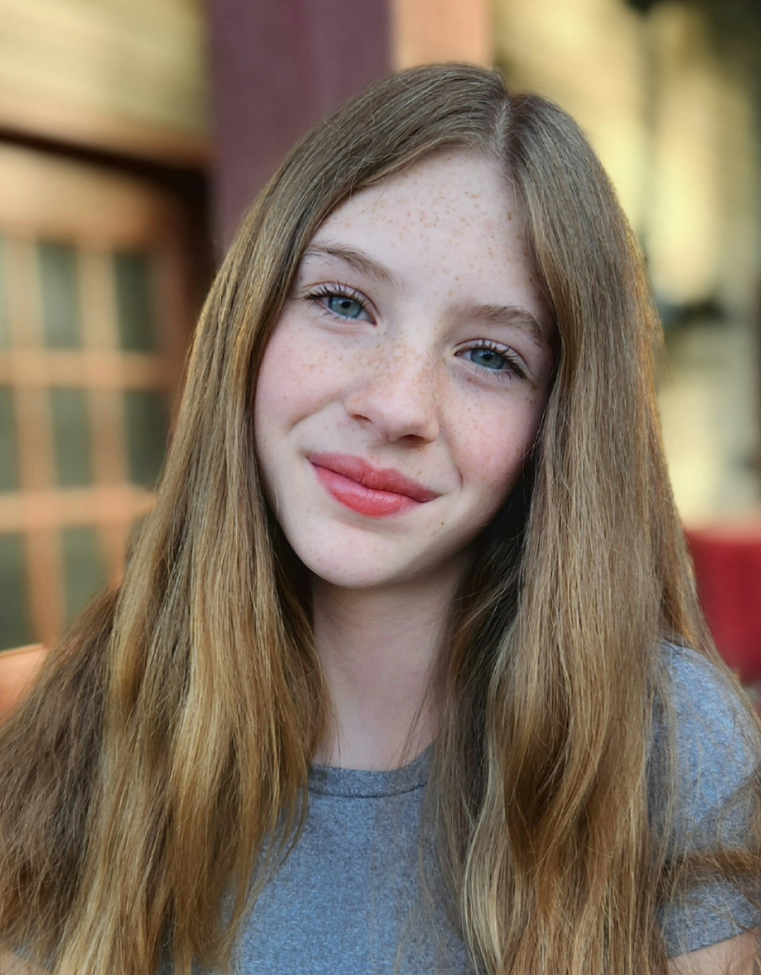 A person with long hair and freckles, wearing a gray shirt, smiling softly with a blurred background.