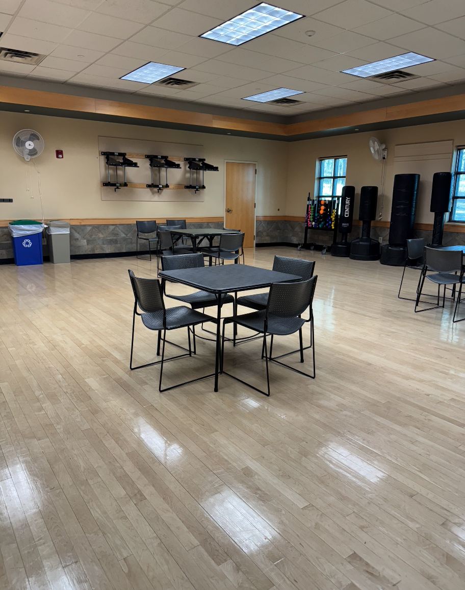 A room with tables, chairs, exercise equipment, fans, and trash bins on a wooden floor.