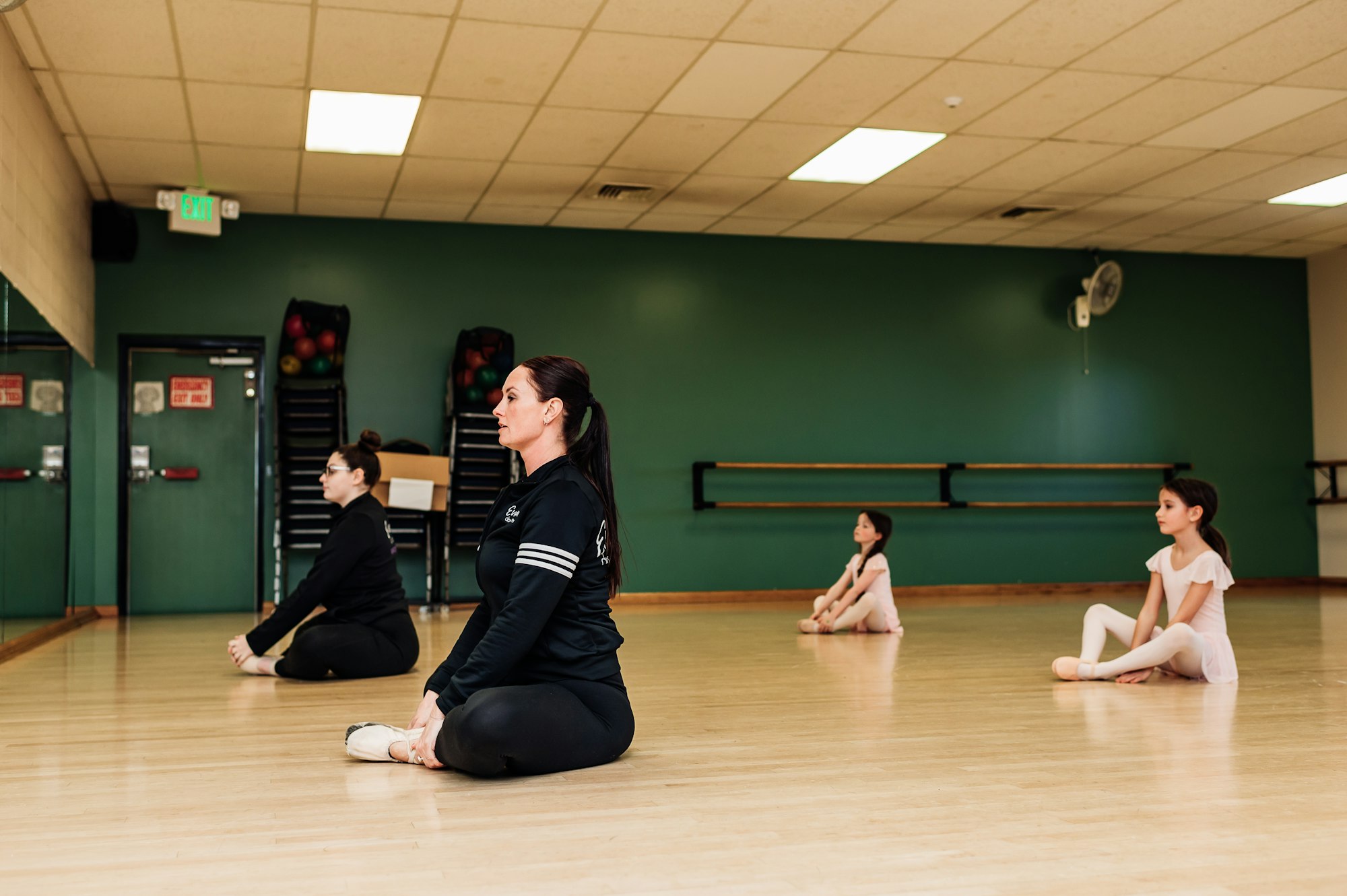 Four people sitting on a dance studio floor in a stretching position, with a green wall and exercise equipment in the background.