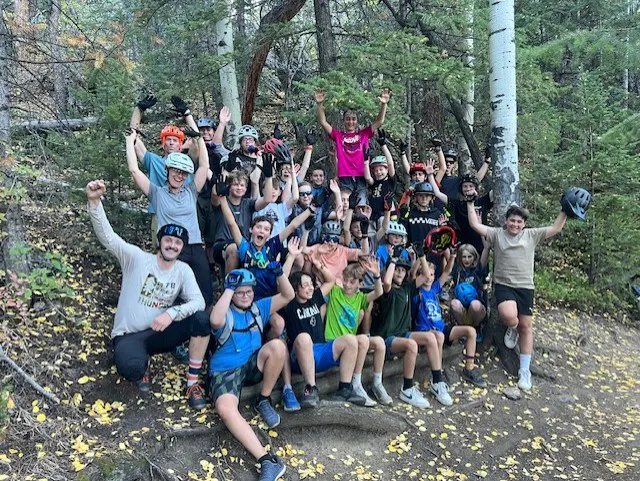 A large group of kids and adults in helmets celebrating outdoors, surrounded by trees and fall foliage.