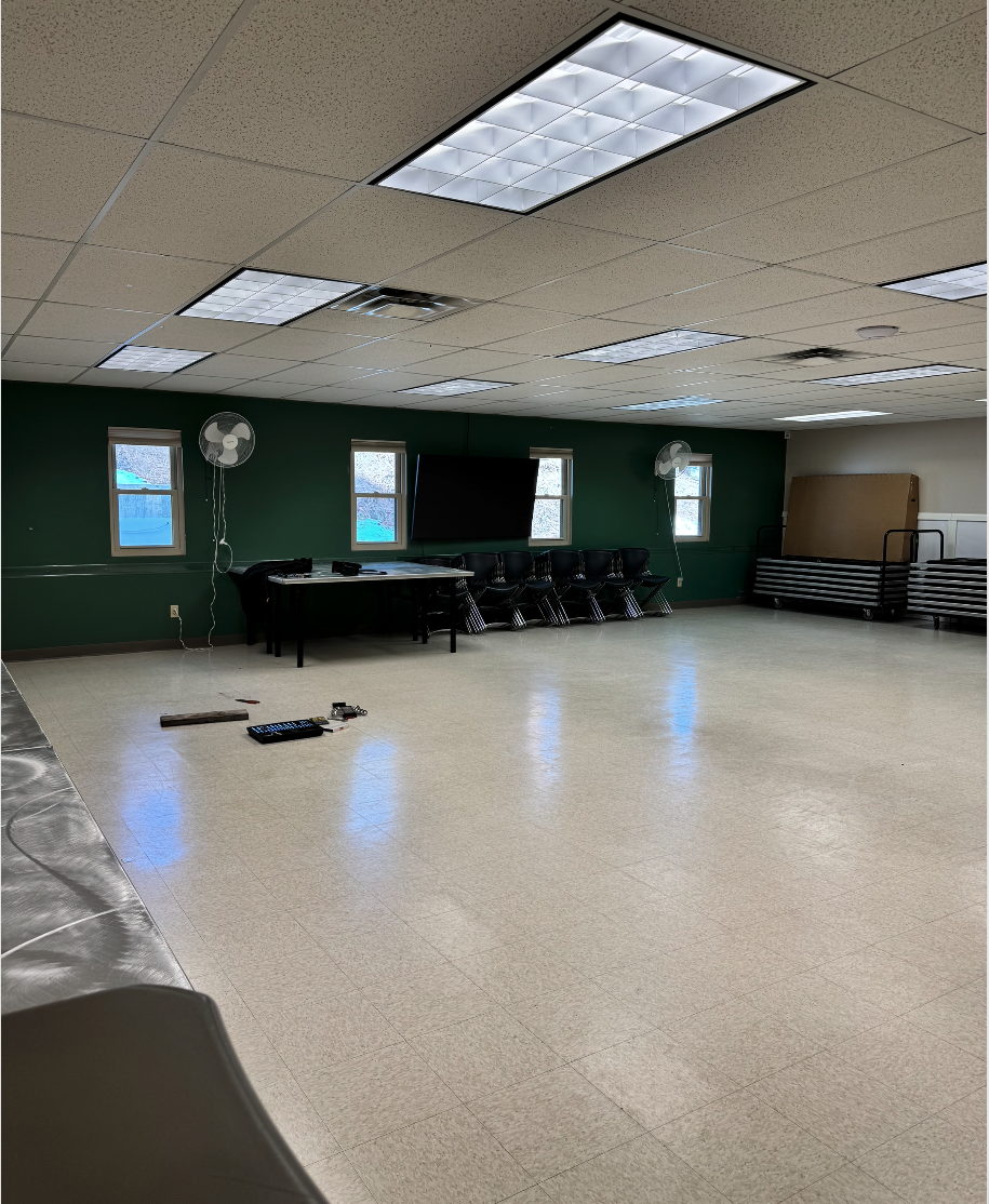 Empty room with tile floor, tables, chairs, fans, a large screen, and some tools on the floor. Ceiling has fluorescent lights.