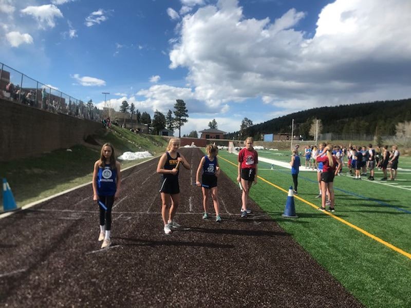 Athletes on a track field, blue skies with clouds, spectators on the side, green hill in the background.
