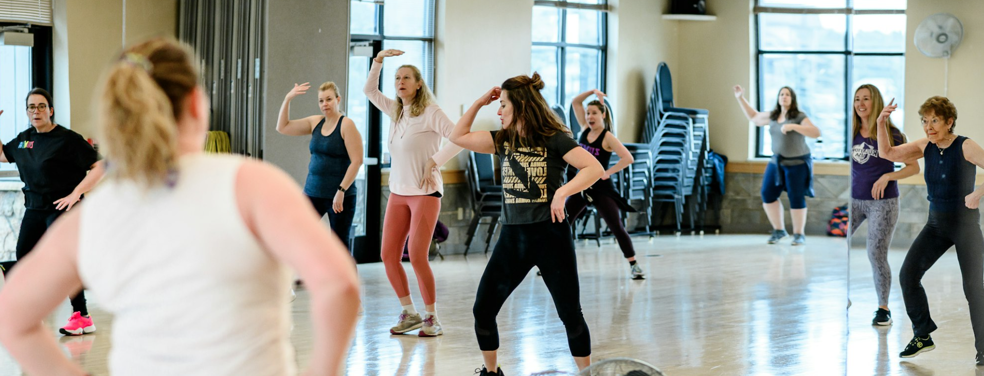A group of people in a gym room participating in a dance or fitness class, following an instructor's movements.